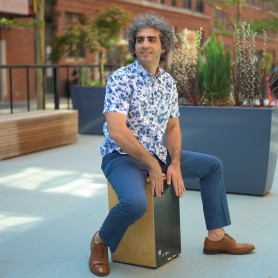 A picture of a man named Tareq Rantisi. He has graying curly hair, is wearing brown slacks, and is sitting in a straddle position atop a wooden block on an outdoor patio in the daytime. He wears a blue and white floral short sleeve button down top with matching solid blue pants. Looking off to the side in curiosity he has his hands resting on the block and his feet pointed outwards.