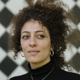 A picture of a woman named Diana Al-Halabi. She has loose curly hair, put up into a frizzy updo. She wears a silver necklace with a black turtleneck, and matching dark eyeliner. She is against an intricate black and white diamond checkered background, faced forward with a resting face but her eyes are wandering to the right curiously.