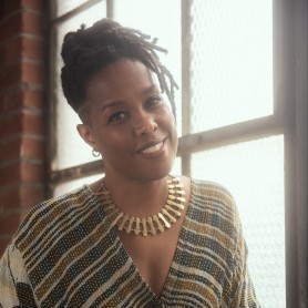 A picture of a genderqueer woman named Laurie Townshend. She is standing with a radiant glow from the window beside her, gently resting an arm on its sill. She wears a striped and patterned v neck dress, with her head cocked to the side and locs styled atop her head. Her neck is adorned with a large gold chain necklace, and she smiles fondly towards the camera.