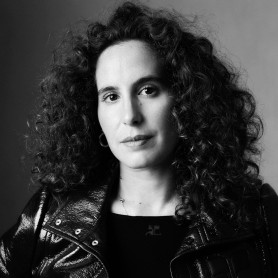 A black-and-white picture of a woman named Lina Soualem. She is faintly smiling at the camera, her curly dark hair against a gradient background. She is wearing a sleek leather jacket and black top.