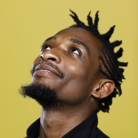 A picture of a man named Nelson Makengo. His head is tilted slightly upwards, and he is happily looking up towards the upper left corner of the image. He is smiling against a solid, yellow color background. He is wearing a high-collared, dark garment. The texture of the skin is clearly visible, with a short, trimmed beard along the jawline and chin, and a mustache. The hair is styled in short twists or locks.