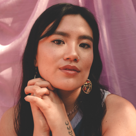 A picture of Jalena Keane-Lee in a serene and contemplative pose against a backdrop of soft, flowing purple fabric. Her long, black hair frames her face, and she is wearing a pair of gold, abstract face-shaped earrings. Her expression is calm and thoughtful, with a slight smile on her lips. She has a nose ring, and her hands are gently clasped under her chin, revealing a small tattoo of moon phases on her forearm.