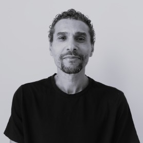 A black-and-white image of a man named Carlos Yuri Ceuninck. He stands against a plain, light-colored background. He has short, tightly curled hair and a neatly trimmed goatee. He is wearing a simple, dark-colored t-shirt with short sleeves. He has a calm and slightly confident expression, with a gentle smile playing on his lips and a relaxed demeanor.