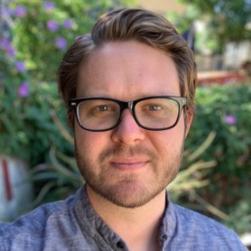 A picture of a man named Lucas Guilkey outdoors. He has light skin, short brown hair styled neatly, and a short beard. He is wearing black rectangular glasses and a button-up shirt in a grayish-blue color. His expression is neutral, with a slight, calm smile, and his eyes looking directly into the camera. The background is blurred but shows a garden with green foliage and purple flowers.