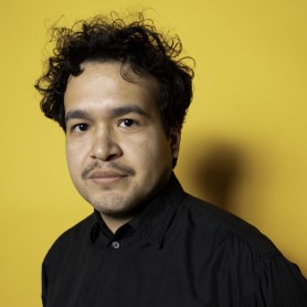A picture of a man named Joebill Munoz standing in front of a plain, bright yellow background. He has dark, curly hair, a mustache, and a short, trimmed beard. He is wearing a black, button-up shirt, and is gazing directly at the camera with a neutral expression.