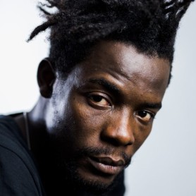 A picture of a man named Pierre Michel Jean against a simple, light gray background. He has a dark complexion, and his hair is styled in multiple short, twisting dreadlocks that are spiked upward and outward. He is wearing a dark-colored shirt, and his facial expression appears serious and contemplative. His eyes are directed towards the camera, with a slight furrowing of the brows and relaxed lips.