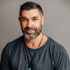 A picture of a man named Damon Fepule'ai against a neutral gray background. He is seated and facing the camera, with a slight smile and warm expression. Damon has short, neatly trimmed hair, and a full beard that is peppered with gray. He is wearing a charcoal gray t-shirt and has a greenstone pendant hanging from a black cord around his neck.