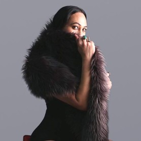 A picture of a transgender person named Ramon Te Wake against a plain gray background. She is dressed in a black, sleeveless bodysuit with a large, plush voluminous fur scarf draped around their shoulders. She holds the fur scarf close with one hand covering her mouth and gazing intently toward the camera, an ornate ring with a prominent green stone adorned on her finger.