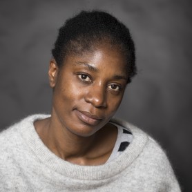 A picture of a woman named Rosine Mbakam, set against a soft-focus gray background. She is positioned slightly off-center, leaning her head gently to the right. She has short tightly coiled black hair, closely-cropped to her head and her expression is calm and introspective, with a soft smile playing on her lips and slightly relaxed eyes. She is wearing a light gray sweater with a wide neckline.
