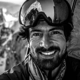 A black-and-white picture of a man named Dominic Gill, smiling at the camera. He has a bushy beard and a slight mustache, and he wears protective ski goggles on his forehead which reflect a mountainous landscape. He is dressed in outdoor gear, including a high-collared jacket. Behind him, the background is slightly out of focus, depicting a dramatic mountain range covered in snow and rugged, jagged peaks.