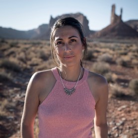 A picture of a woman named Nadia Gill, standing outdoors in a desert landscape. She is centered in the frame, looking directly at the camera with a calm and neutral expression. Nadia has dark brown hair tied back in a simple ponytail. She is wearing a sleeveless, light pink knit top and a distinctive silver necklace composed of fan-like elements hanging in a semi-circle around her neck.