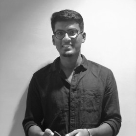 A black-and-white picture of a young man named Shiva Krish standing against a plain white background. He is captured from the waist up, wearing a dark, long-sleeved button-up shirt with the sleeves rolled up to his elbows. He has short, neatly styled hair and wears round glasses. He has a slight beard and a friendly smile with his hands positioned in front of him, holding a mobile phone.