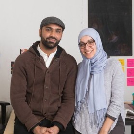 A picture of two individuals. Turab Shah, a man on the left, and Arwa Aburawe, a woman on the right, stand closely together in an office environment. Turab Shah is dressed in a brown cardigan over a white shirt and wears a flat cap. He has a neutral expression with a hint of a smile. Arwa Aburawe is wearing a light gray hijab, glasses, and a matching light gray top. She has a broad, warm smile. The background features a cluttered bulletin board filled with various colorful notes, postcards, and photos.
