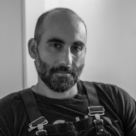 A black-and-white picture of a man named Andy Sarjahani. He is facing the camera directly and has a shaved head and a full beard peppered with gray. He is wearing dark-colored overalls over a dark shirt against a solid light background.