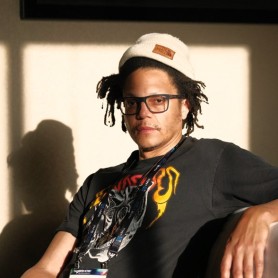 A picture of a man named Raafi Rivero sitting casually in a sunlit room. The light casts a prominent shadow of him on the beige wall behind. He is wearing a white beanie with a brown leather patch, large rectangular glasses, and a black T-shirt featuring vibrant yellow and red graphics. He has medium-length dreadlocks that extend from under his beanie.