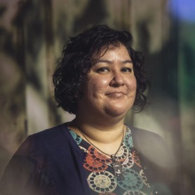 A picture of a woman named Erika Valenciana, standing against an out-of-focus background with a mix of light and shadow. She has short, curly dark hair and is wearing a thoughtful expression with a slight smile. She is dressed in a colorful shirt adorned with circular patterns resembling film reels, featuring hues of orange, teal, and pink. Over her shirt, she wears a navy-blue cardigan. Around her neck, Erika has a silver necklace with a prominent pendant shaped like a hand.
