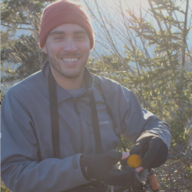 A picture of a man named Mitch Wenkus. He is in a natural setting with evergreen trees in the background, under a bright, sunny sky casting a light glow. He smiles warmly and is wearing a red beanie, a gray jacket, and black gloves. He is holding a camera that he has around his neck.