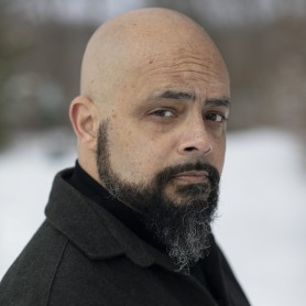 A picture of a man named Robe Imbriano set against a blurred snowy background. He is turned slightly to his left, and looking directly at the camera with a pensive and introspective expression. He is bald with a full, neatly trimmed beard that is speckled with gray. He is wearing a dark gray, woolen coat over a black turtleneck.