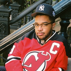 A picture of a man named Ché Williams sitting on an outdoor staircase. He is wearing a black New York Yankees baseball cap and a red New Jersey Devils hockey jersey, marked with a large white "C" on the right side. He has glasses and a short beard. His hands are clasped together resting on his lap, and he has a calm, composed expression on his face.