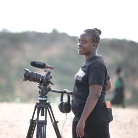 A picture of a woman named Laissa Malih, standing outdoors with a professional camera mounted on a tripod. Laissa is smiling and looking slightly over her shoulder at the camera. She has braided hair tied back and is wearing a short-sleeved black t-shirt and black pants. The background is a blurred, natural landscape with some greenery and distant hills.