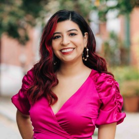 A picture of a non-binary person named Kristal Sotomayor standing outdoors. She is dressed in a vibrant fuchsia wrap dress that features puffed sleeves and a belt tied at the waist. Her dark hair, which has a red highlight on one side, falls in soft waves over her shoulders. She is smiling broadly with a friendly expression.
