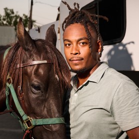 A picture of a man named Drake LeBlanc standing closely beside a brown horse. He has short, styled dreadlocks and a neatly trimmed beard and wears a light gray polo shirt, dark pants, and a confident expression. The horse looks serene with its ears pointed upwards and its tongue slightly sticking out.