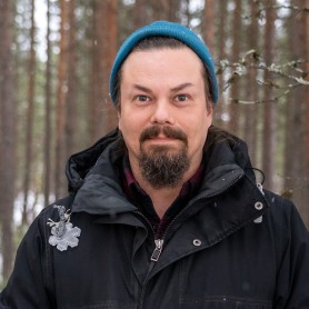 A picture of a man named Tuomas Kumpulainen standing in a forest. He is wearing a teal beanie, a dark jacket with a high collar, and a decorative silver brooch shaped like a snowflake pinned to the left side of his jacket. He has long dark hair tucked behind his ears, a mustache, and a goatee. His expression is neutral but engaged, with wide eyes looking directly at the camera.