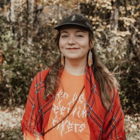 A picture of a non-binary person named Sunná Nousuniemi, standing outdoors against a backdrop of dense forest with trees displaying autumn foliage in shades of yellow and green. Sunná wears a confident, gentle smile, has light skin, and long brown hair styled in two low braids. They wear a black cap with white embroidery, a pair of long, multi-colored beaded earrings, and a red shawl with a tartan pattern draped over their shoulders.