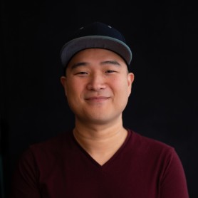 A picture of a man named John Beder against a black background. He is centered in the frame and looking directly at the camera with a gentle smile. John is wearing a dark gray baseball cap with a light gray brim and a maroon V-neck T-shirt.