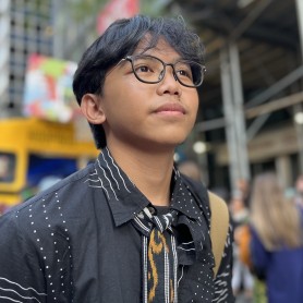 A picture of a young man named Kynan Tegar, who is centered in the frame and appears to look slightly upwards with a serene expression. He has short, neatly trimmed black hair, and wears rectangular-framed glasses. He is dressed in a black shirt adorned with white dotted and lined patterns, and a tie with a black, white, and gold design around their neck. The background is slightly out of focus, showing a vibrant street scene with a crowd of people.