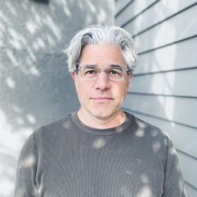 picture of a man named Ross Kauffman. He has gray hair and clear-framed glasses. He appears to be middle-aged, with a calm and neutral expression on his face. He is wearing a gray, long-sleeved, textured shirt.