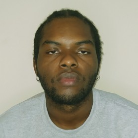 A picture of a man named Andres Mosquera. He is pictured from the chest up against a plain, light-colored background. He has a serious expression and is looking directly at the camera. Andres has a medium-length afro hairstyle, with closely cropped facial hair outlining his jaw. He is wearing a light gray sweatshirt.