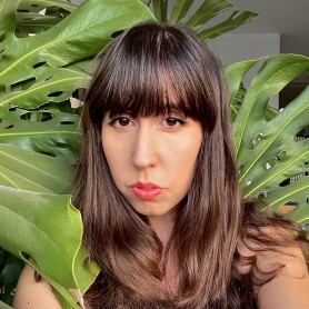A picture of a woman named Tiare Ribeaux. She has long, slightly wavy brown hair with bangs that fall just below her eyebrows. Her expression is serious, with neutral lips accented by red lipstick, and her eyes feature dark eyeliner. She is surrounded by large, vibrant green Monstera leaves with distinctive holes and perforations.