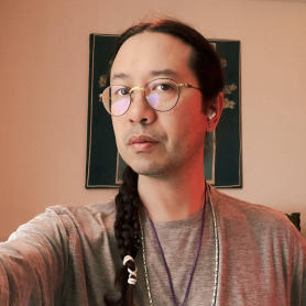 A picture of a man named Jody Stillwater. They are indoors, standing in front of a light pink wall with an ornate, green, and gold framed artwork hanging on it. He has long, dark hair tied back in a braid adorned with white beads and is wearing round, gold-rimmed glasses. He is dressed in a simple gray long-sleeve shirt and is accessorized with multiple thin necklaces, and he has a neutral and calm demeanor.