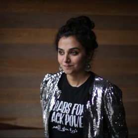 A picture of a woman named Alia Haju. She is standing indoors against a background of wooden planks with alternating shades of brown. Alia has dark, curly hair pulled up into a bun and wears large, intricate silver earrings. She is dressed in a shiny, silver sequin jacket over a black T-shirt that reads "ARABS FOR BLACK POWER" in bold white letters, along with a line of Arabic text beneath.
