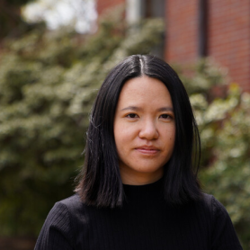 A picture of a woman named Bea Hesselbart. She is standing outdoors with a softly blurred background of greenery and a red brick building. Bea has shoulder-length straight black hair and is wearing a black top that blends seamlessly with her dark hair. Her expression is calm and composed, with a faint, gentle smile.