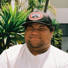 A picture of a man named Laman Time. He is outdoors and is positioned in front of a background filled with large, green, spiky leaves of tropical plants. He is wearing a white T-shirt and a black baseball cap with an emblem that reads "Stampede City Calgary, Alta." The man has a neutral expression, with his eyes squinting.