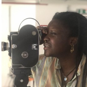 A picture of a woman named Simisolaoluwa Akande, who is shown closely examining a vintage film camera mounted on a tripod. She has a focused expression, with one eye closed as she looks through the camera’s viewfinder. She is wearing an open-collared, pastel-colored shirt with patterns, and an ornate earring hangs from her ear.