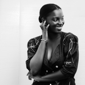 A black-and-white picture of a woman named Maame Adjei. She stands against a plain white background, wearing a dark long-sleeved blouse with intricate, light-colored line patterns and high-waisted black pants. The blouse has a deep V-neck and is adorned with buttons. Her hair is styled in a low, neat bun. She has her left hand resting on her right arm, and her right hand gently touching her face while smiling warmly.