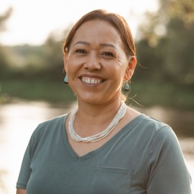 A picture of a woman named Katsitsionni Fox standing outdoors. She is depicted smiling warmly with a gentle expression. She has medium-length hair that is tied back, and she wears a pair of turquoise earrings. She is dressed in a light gray V-neck shirt with a small pocket on the left side. Around her neck is a beaded necklace of a lighter hue. The background is softly blurred, revealing a lush greenery and a body of water.