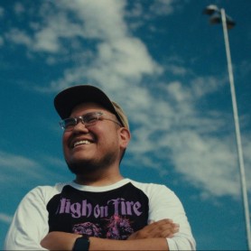 A picture of a man named Fritz Bitsoie standing outdoors against a vibrant blue sky filled with scattered white clouds. He is smiling broadly, showing a cheerful and relaxed expression. He is wearing a light tan baseball cap and clear-framed glasses. His shirt is black with white sleeves, and the front displays the text “High on Fire” in bold, stylized purple letters, accompanied by intricate artwork and his arms are crossed over his chest.