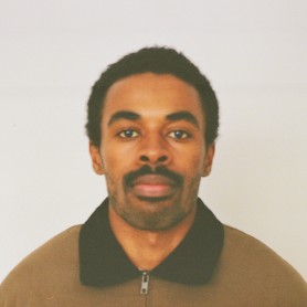 A picture of a man named Blair Barnes. He is positioned against a plain, light-colored background and has short, curly hair and a neatly trimmed mustache. He is wearing a brown zip-up jacket with a black collar. His expression is neutral, with a calm and composed demeanor.