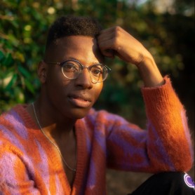 A picture of a man named Michael Donte. He is seated outdoors in a sunlit setting. He is wearing round, clear glasses and a distinctive sweater with bold, striped patterns of bright orange and lavender shades. His expression is contemplative as he gently rests his right hand on his head.