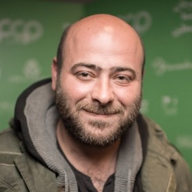 A picture of a man named Nidal Badarny. He has a bald head and a neatly trimmed beard with a friendly, subtle smile that suggests warmth. He is wearing a dark gray jacket layered under a green coat, both of which look slightly worn and casual, suitable for cool weather. The blurred background is a green backdrop with logos and text.