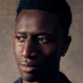 A picture of a man named Welket Bungué. He has short, curly black hair and is wearing a dark, pinstriped jacket. His expression is contemplative as he gazes off to the right. A small, simple hoop earring is visible on his left ear.