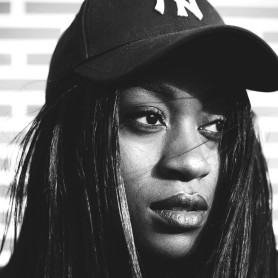 A black-and-white picture of a woman named Victoria Neto. She is captured from the shoulders up with a strong and resolute expression. She is wearing a black baseball cap featuring an embroidered “NY” logo. Her long, straight hair cascades down around her face, partially obscuring her right eye.