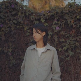 A picture of a woman named Joanne Mony Park. She stands with her hands in her pockets, against a backdrop of dense, vine-covered foliage with patches of green and some scattered violet flowers. She is looking off to the side with a contemplative expression. She has medium-length, dark hair that frames her face and wears a light gray, oversized button-up shirt over a white top.