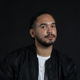 A picture of a man named Joseph Douglas Elmhirst against a dark background. He has a composed expression with a subtle smile on his face, eyes looking slightly to the side. His hair is styled in neatly braided cornrows. Joseph is wearing a black bomber jacket over a white t-shirt. Around his neck, he has a delicate gold chain with a small cross pendant.