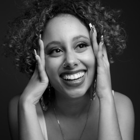 A black-and-white picture of a woman named Imani Celeste. She is wearing a tank top, has curly hair, and her head and eyes are tilted slightly upward. She is smiling widely, displaying a set of straight teeth. Her hands are resting on either side of her face, with long nails and she is wearing large hoop earrings, a nose ring on her right nostril, and a small stud on her left nostril.