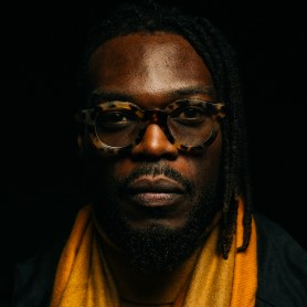 A picture of a man named Walé Oyéjidé. He has a composed expression and is wearing a pair of large, round, tortoiseshell glasses with thick frames. His eyes are partially visible through the glasses, gazing directly at the camera. He has a neatly trimmed beard and mustache, with his hair styled in short dreadlocks, some of which are draped over his shoulder. He is wearing a mustard-yellow scarf wrapped around his neck over a matching turtleneck sweater.